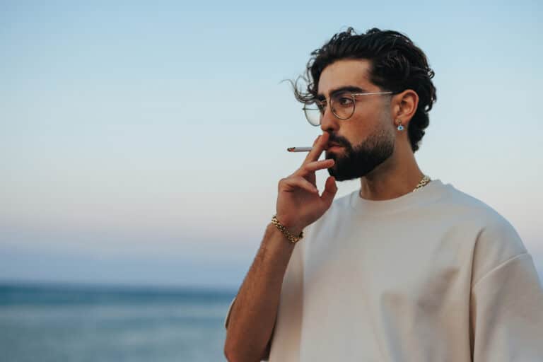 A young cool guy is at the beach at sunset smoking a joint.